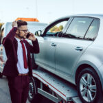 Man on phone standing next to car being loaded onto flatbed before calling a tow truck