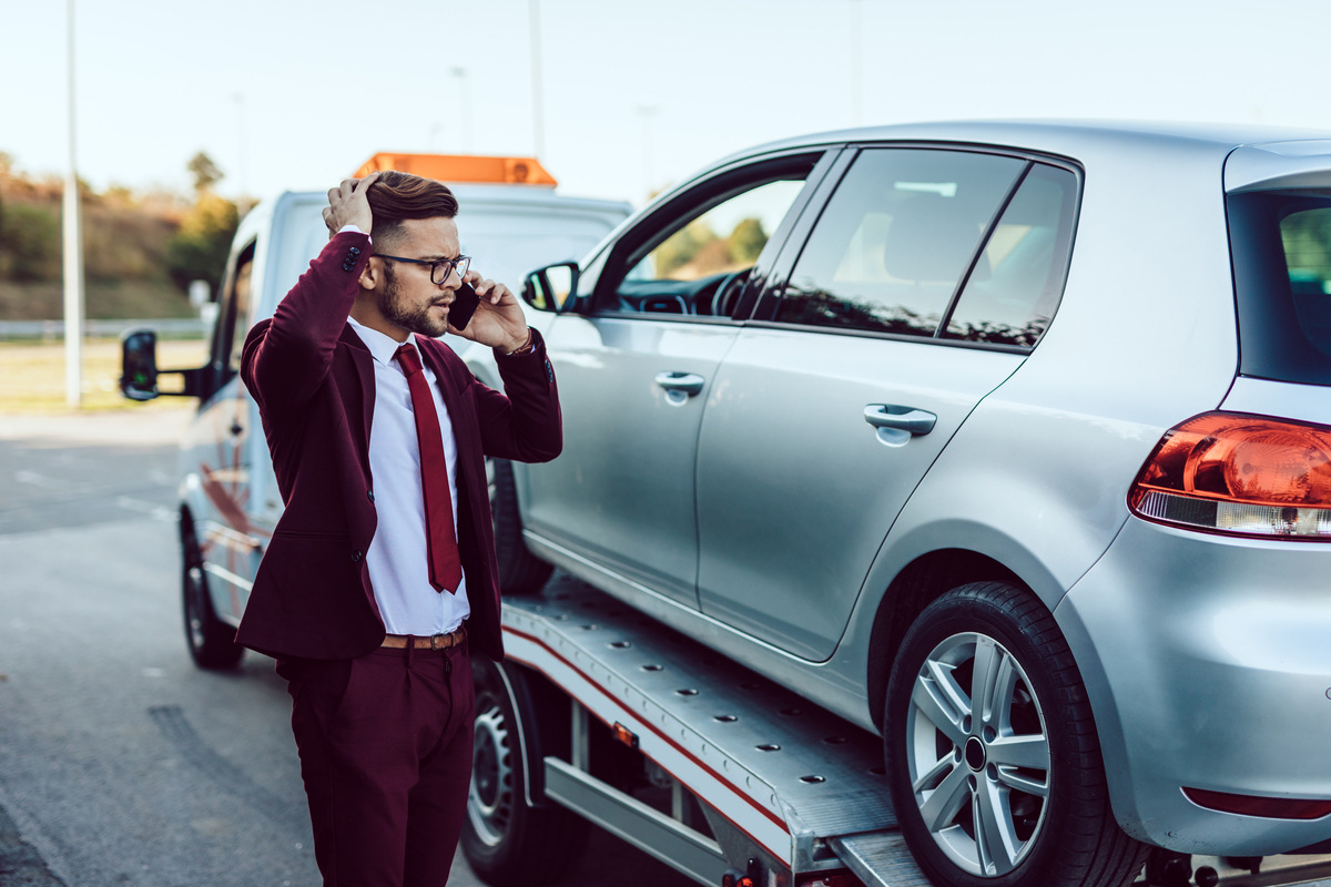 Man on phone standing next to car being loaded onto flatbed before calling a tow truck